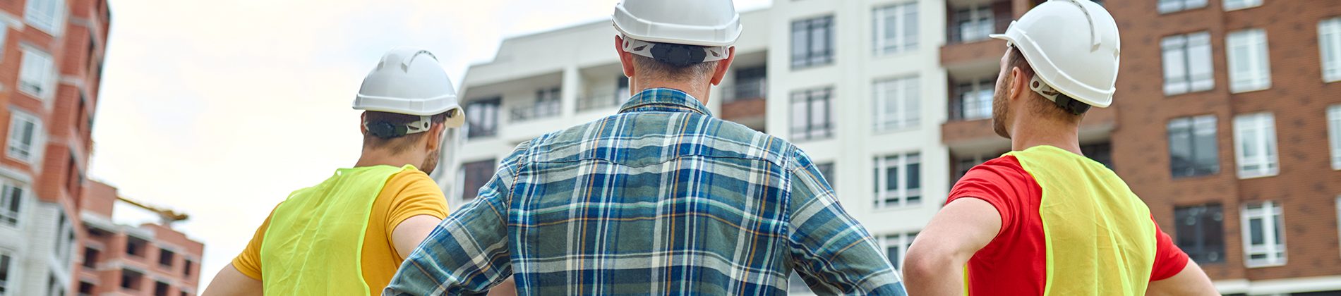 result-back-view-of-three-men-in-protective-helmets-holding-hands-on-his-waist-standing-at-construction-site-looking-at-buildings-under-construction-during-day